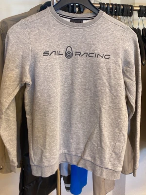 Grå sweatshirt från Sail Racing - Snygg grå sweatshirt från Sail Racing med svart logotyptryck på bröstet och liten logga i nacken. Tröjan har rund halsringning, långa ärmar och är tillverkad i mjukt material. Perfekt för en avslappnad och sportig stil.