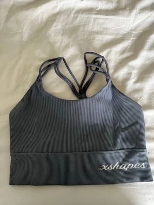 Blå sport-bh från xshapes - Snygg grå sport-bh från xshapes med ribbat tyg och dubbla smala axelband som korsas och flätas i ryggen. Perfekt för träning och har en stilren design med logga framtill. Materialet är stretchigt och bekvämt.
