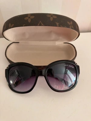 Svarta Louis Vuitton solglasögon - Snygga svarta solglasögon från Louis Vuitton med stora, rundade bågar och mörka glas som tonar mot lila. Kommer med klassiskt LV-fodral i brunt med monogrammönster. Perfekta för dig som vill ha en lyxig och trendig accessoar.