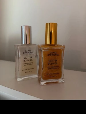Gamma Aromatics Glitter Body Oil - Två flaskor Glitter Body Oil från Gamma Aromatics. En med guldigt glitter och guldfärgat lock, en med silvrigt glitter och silverfärgat lock. Innehåller kokosolja och vitamin E. Passar alla hudtyper och ger huden en skimrande finish.