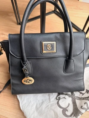 Svart handväska från Christian Lacroix - Stilren svart handväska från Christian Lacroix i strukturerat konstläder. Väskan har två handtag, avtagbar axelrem och guldfärgade metalldetaljer, inklusive logoplatta och hänge. Klassisk fyrkantig form och dragkedja upptill.