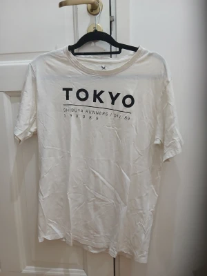 Vit T-shirt med Tokyo-tryck - Vit T-shirt med svart text 'TOKYO' och 'Shibuya Runners / Div. 89' framtill. Klassisk rund hals och korta ärmar i bommulls material