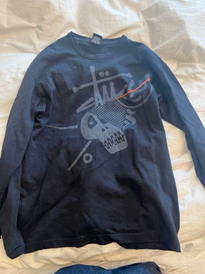 Svart långärmad tröja från Stüssy - Svart långärmad tröja från Stüssy med stort tryck framtill som inkluderar en dödskalle och grafiska detaljer i grått och rött. Tröjan har rund hals och är tillverkad i mjuk bomull, perfekt för en streetwear-look.