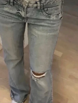 Ljusblå slitna bootcut jeans - Säljer ett par ljusblå bootcut jeans med slitning och hål på ena knät. Jeansen har klassisk femficksmodell och låg midja. Perfekta för en avslappnad och trendig look. Tillverkade i denim med skön passform.