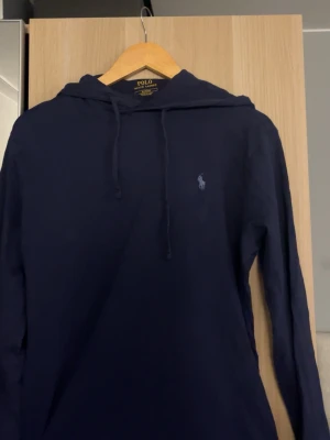 Mörkblå hoodie från Polo Ralph Lauren - Snygg mörkblå hoodie från Polo Ralph Lauren med klassisk huva och dragsko. Ikoniska broderade loggan på bröstet. Tillverkad i mjukt material som är skönt att bära. Perfekt för en avslappnad och stilren look.