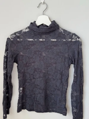 Mörkgrå spets polotopp - Snygg mörkgrå polotopp i genombruten spets med blommigt mönster. Långärmad och tight passform, perfekt för att styla lager på lager eller som statement piece. Tunn och stretchig, ger en cool och elegant vibe.