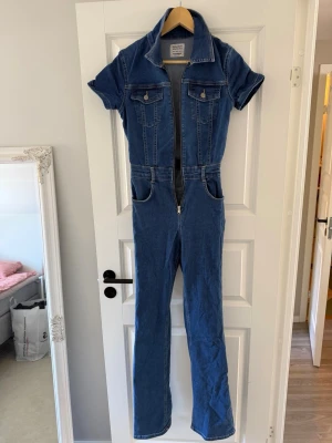 Blå denim jumpsuit med kort ärm från Pull&Bear - Snygg blå denim jumpsuit från Pull&Bear med korta ärmar, krage och dragkedja framtill. Två bröstfickor med lock och två sidfickor. Figurnära passform upptill och utsvängda ben. Perfekt för en cool och avslappnad look.