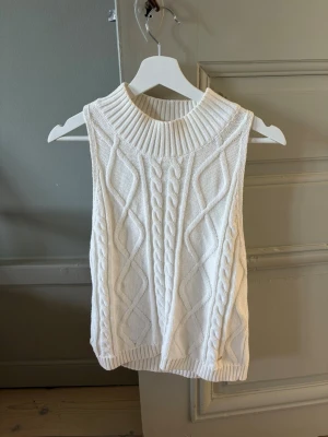 Vit kabelstickad ärmlös tröja - Säljer en vit kabelstickad ärmlös tröja med hög ribbad krage. Tröjan har ett klassiskt flätmönster framtill och ribbade kanter nedtill. Perfekt att styla över en skjorta eller långärmad topp för en trendig lager-på-lager-look.