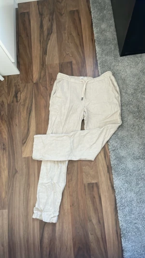 Beige linnebyxor  - Säljer ett par ljusbeige linnebyxor med dragsko i midjan och avslappnad passform. Byxorna har resår i midjan och muddar vid bensluten samt sidofickor. Perfekta för en chill och stilren look.