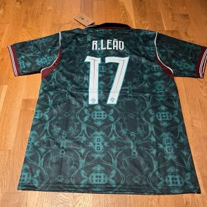 Portugal green R.Leao #17 - Snygg Portugal fotbollströja från Raske med R. Leão och nummer 17 på ryggen. Tröjan är mörkgrön med turkost mönster, vinröda detaljer på sidor och ärmslut samt vit krage. Perfekt för dig som älskar fotboll och vill sticka ut på planen.