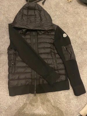 Svart pufferjacka från Moncler - Snygg svart pufferjacka från Moncler med stickade ärmar och huva.Storlek S Jackan har dragkedja framtill, två sidofickor med dragkedja och en extra ficka med dragkedja på ärmen. Moncler-logga på vänster ärm. Jackan är endast andvänd ett fåtal gånger och är i ett superbra skick. Priset början från 2000 men det priset går att diskuteras ända ner till 1400 beroende på hur snabb du är!
