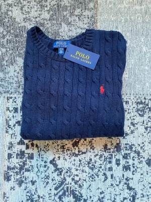 Ralph Lauren kabelstickad tröja marinblå (14–16 / XS) - Äkta Ralph Lauren kabelstickad tröja i klassisk marinblå färg med röd logotyp. Storlek L / 14–16 år. Passar även XS dam beroende på önskad passform. Köpt på KidsBrandStore och i mycket fint skick, tröjan är  använd en gång. Tidlös och populär modell. Skriv gärna vid frågor eller intresse 😊