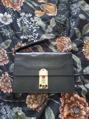 Vintage handloväska / "mansväska" i svart läder – Klassisk modell - Säljes:  Klassisk vintage handloväska (handledsväska) i tidlös svart design. Väskan är tillverkad i läder och har ett dekorativt kombinationslås i guldfärgad metall på framsidan. En perfekt accessoar för den som gillar 80- eller 90-talsstil, eller behöver en smidig väska för värdesaker och dokument. Utsida: Svart läder med snygga stickningar och en praktisk handledsrem. På baksidan finns ett extra fack med dragkedja. Insida: Väskan är rymlig med flera fack, varav ett med dragkedja för säker förv