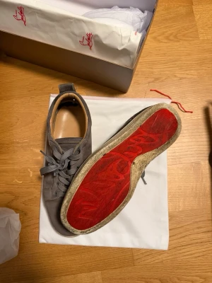 Gråa Louboutin sneakers med röd sula - Snygga gråa sneakers från Christian Louboutin i mocka med klassisk röd sula. Skorna har lågt skaft, rund tå och gråa skosnören. Perfekta för dig som vill sticka ut med exklusiva detaljer och ikonisk design.