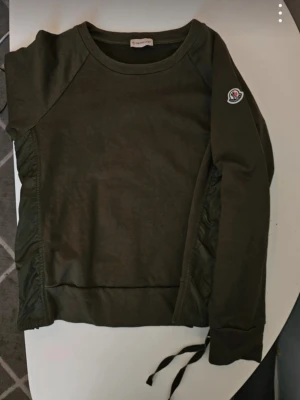 Olivgrön sweatshirt från Moncler - Cool orginal olivgrön sweatshirt från Moncler med rund halsringning och långa ärmar. Tröjan har quiltade detaljer längs sidorna och en dragsko nertill. Moncler-logga på ena ärmen ger en exklusiv touch. Perfekt för en avslappnad men stilren look.