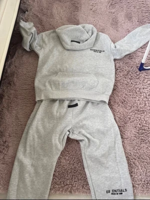 Grå mjukisbyxor + hoodie från Essentials - Snygga grå mjukisbyxor från Essentials Fear of God med svart logga på benet. Byxorna har resår i midjan och är gjorda i mjukt bomullsmaterial för maximal komfort. Perfekta för en chill och avslappnad stil.