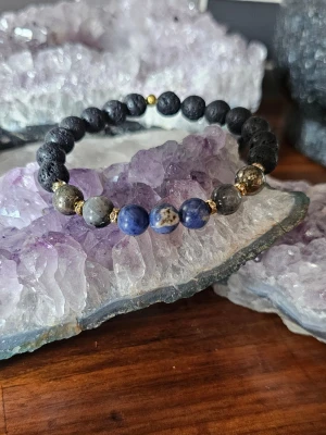 Armband med svarta och blå pärlor - Stilrent armband med halvädelstenar som  lavasten, Sodalit, larvikit och pyrit, dekorerat med små guldfärgade detaljer mellan pärlorna. Elastisk tråd gör det enkelt att ta på och av. Perfekt accessoar för att lyfta din outfit. Det levereras i en söt liten påse tillsammans med information om kristallernas magiska egenskaper. 