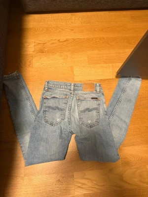 Ljusblå jeans från Nudie Jeans - Säljer ett par ljusblå jeans från Nudie Jeans med klassisk femficksdesign och tydliga sömmar på bakfickorna. Jeansen har raka ben och är tillverkade i bomull. Perfekta för en avslappnad och trendig stil.