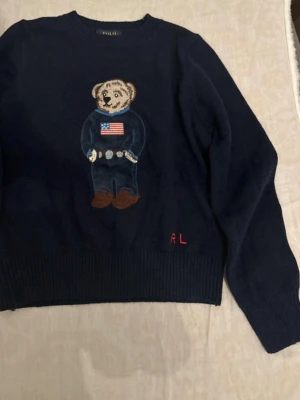 Mörkblå stickad tröja Polo Bear - Mörkblå stickad tröja från Ralph Lauren med ikoniska Polo Bear broderad framtill. Tröjan har rund hals, långa ärmar och ribbade muddar. Detaljerad björn med amerikansk flagga på tröjan och RL-broderi nertill. Perfekt för dig som gillar klassisk preppy stil.