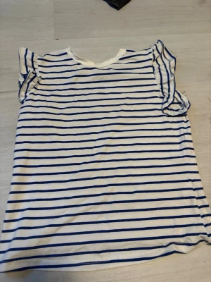 Randig vit och blå topp med volangärm - Vit topp med blåa horisontella ränder och volangdetaljer på ärmarna. T-shirten har en avslappnad passform och är perfekt för en fräsch och somrig look. Tillverkad i mjukt bomullsmaterial.tveka inte att höra av er vid frågor eller funderingar😇💕