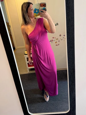 Lila oneshoulder långklänning - Snygg lila långklänning med oneshoulder-design och draperad detalj vid midjan. Klänningen har en smickrande passform och är tillverkad i ett mjukt, följsamt material som ger en elegant siluett.Helt. Ny