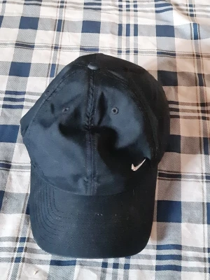 Svart Nike keps med broderad logo - Snygg svart keps från Nike med klassisk böjd skärm och liten vit broderad swoosh-logo framtill. Kepsen har ventilerande hål och justerbar rem baktill. Perfekt accessoar för en sportig och avslappnad stil.