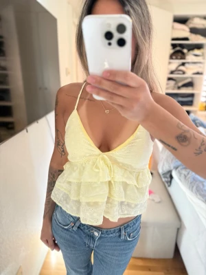 Gul volang topp med rosett - Söt gul linnetopp med tunna axelband, volanger och en stor rosett framtill. Toppen har broderade detaljer och är croppad, perfekt för varma dagar. Passar dig som gillar en feminin och lekfull stil. Står ingen storlek passa mig som bruka ha xs-s