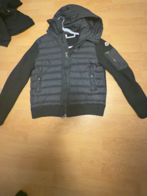 Svart pufferjacka från Moncler - Snygg svart pufferjacka från Moncler med stickade ärmar och huva. Jackan har dragkedja framtill, två fickor med knappar och en extra ficka på ärmen. Materialmix av dunfylld kropp och stickade detaljer ger en cool look.