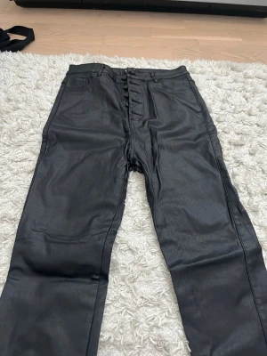 Svarta coated jeans från Rock Angel - Snygga svarta coated jeans från Rock Angel med hög midja och knäppning framtill med stora svarta knappar. Jeansen har klassisk femficksdesign och en glansig finish som ger en cool look. Perfekta för dig som vill sticka ut lite extra.