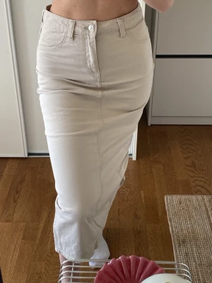 Beige långkjol i denim - Snygg beige långkjol i denim med klassisk femficksmodell och hög midja. Kjolen har en rak passform och stängs med knapp och dragkedja framtill. 