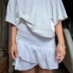 Vit kortkjol med volang - Supersöt vit kortkjol med volangdetalj som ger en extra touch. Det är en omlottkjol i jeans tyg. Perfekt för dig som vill ha en fräsch och enkel look till sommaren.