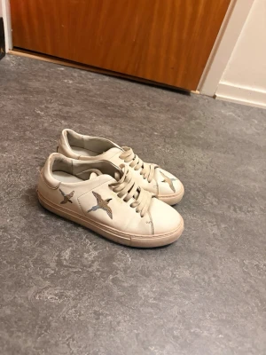 Vita Axel Arigato sneakers med broderade fåglar  - Snygga vita sneakers med broderade fåglar i blått och brunt på sidorna och framtill. Skorna har klassisk låg modell med vita snören och rund tå. Tillverkade i slätt material som ger en clean look. Perfekta för dig som vill ha något unikt och stilrent. Säljer då dessa skor för att de är små på mig