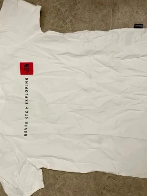 Vit t-shirt från The North Face - Vit t-shirt från The North Face med rött logomärke på bröstet och texten 'Never Stop Exploring' tryckt vertikalt. Klassisk passform och korta ärmar, perfekt för en avslappnad stil.