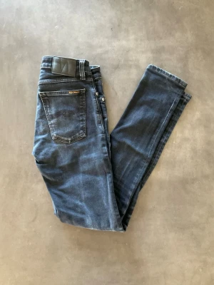 Mörkblå jeans från Nudie Jeans - Säljer ett par mörkblå jeans från Nudie Jeans med klassisk femficksmodell och smal passform. Jeansen har en snygg tvätt och detaljerad söm på bakfickan samt svart patch i midjan. Perfekta för dig som gillar stilrena och tidlösa jeans.
