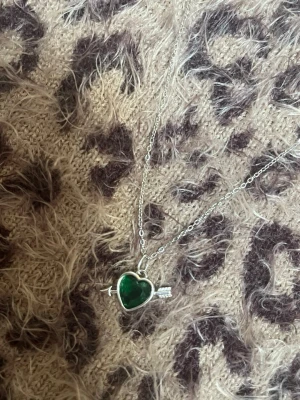 Halsband med grönt hjärthänge silver - Snyggt halsband i silverfärgad metall med ett hjärtformat hänge i grön sten. Hänget har en pil genom sig vilket ger en cool och lekfull vibe. Perfekt accessoar för att lyfta din outfit med en färgklick och unik detalj. Rostfri 