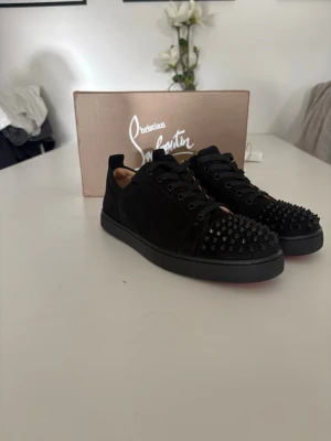 Christian Louboutin svarta sneakers - Svarta sneakers från Christian Louboutin i mocka med svarta nitar på tån och klassisk röd sula. Skorna har lågt skaft, svarta skosnören och en stilren design som sticker ut med sina detaljer. Perfekta för dig som vill ha något unikt och edgy. Skorna är i perfekt nyskick har aldrig använt de bara testat!