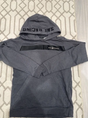Grå hoodie från Sail Racing - Mörkgrå hoodie från Sail Racing med svart detalj och logga på bröstet samt text på huvan. Klassisk känguruficka framtill och mjukt bomullsmaterial. Perfekt för en avslappnad och sportig stil.