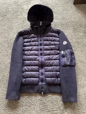 Mörkblå pufferjacka från Moncler - Snygg mörkblå pufferjacka från Moncler med stickade ärmar och vadderad framsida. Jackan har huva, dragkedja framtill och en praktisk ficka med dragkedja på ena ärmen. Moncler-logga på vänster ärm. Perfekt för sommarkvällar!