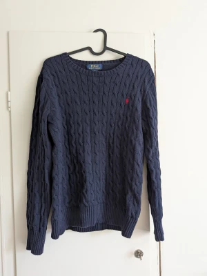 Mörkblå kabelstickad tröja Polo Ralph Lauren - Kabelstickad mörkblå tröja från Polo Ralph Lauren med rund halsringning och lång ärm. Tröjan har ett litet rött broderat logomärke på bröstet och ribbstickade muddar vid ärmslut och nederkant. Perfekt för en stilren och klassisk look.