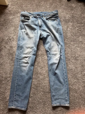 Ljusblå raka jeans från Levi's - Säljer ett par klassiska ljusblå jeans från Levi's med raka ben och fem fickor. Jeansen har en snygg tvätt med slitningar framtill och den ikoniska läderlappen bak i midjan. Perfekta för en avslappnad och trendig stil.