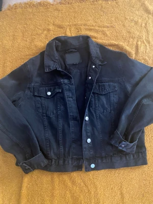 Svart jeansjacka med silverknappar - Stilren svart jeansjacka med klassisk krage och silverfärgade knappar. Två bröstfickor med lock och knapp, samt raka ärmar med knapp vid mudden. Perfekt oversized passform för en cool och avslappnad look.