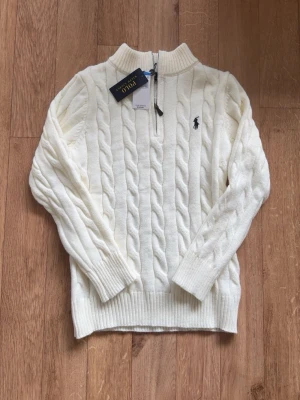 Vit kabelstickad tröja Polo Ralph Lauren - Kabelstickad vit tröja från Polo Ralph Lauren med halv dragkedja och hög krage. Tröjan har ribbstickade muddar och nederkant samt den klassiska broderade loggan på bröstet. Perfekt för en stilren och tidlös look.