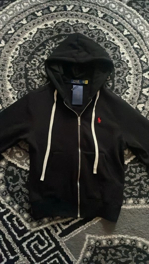 Svart hoodie från Polo Ralph Lauren - Svart hoodie med dragkedja från Polo Ralph Lauren. Tröjan har vit dragsko i huvan, två fickor fram och den klassiska röda logotypen broderad på bröstet. Tillverkad i mjukt bomullsmaterial för en bekväm känsla. Öppen för prisförslag!