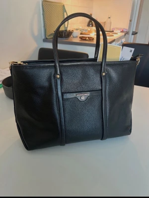 Svart handväska från Michael Kors - Stilren svart handväska från Michael Kors i präglat läder med guldfärgade detaljer. Väskan har dubbla handtag, en ytterficka med knapp och dragkedja upptill. Perfekt för dig som vill ha en klassisk och rymlig väska med exklusiv känsla.
