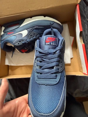 Nike Air Max 90 blå/vit/röd sneakers - Nike Air Max 90 sneakers i mörkblått med vita detaljer och röda Air Max-loggor. Säljer dessa då jag valde fel storlek. Dom är helt oanvända och i helt perfekt skick. Bara att höra av er vid frågor.