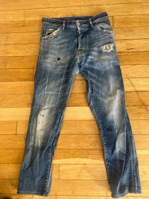 Dsquared2 blå slitna jeans - Coola blå jeans från Dsquared2 med slitningar, hål och färgstänk för en edgy look. Jeansen har klassisk femficksmodell och synliga detaljer som ger en unik streetkänsla. Perfekta för dig som gillar en avslappnad och trendig stil. Storleken passar någon som är mellan 168-170, midja 29/28 skräddarsydda ny pris 6000