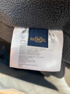  Henry Lloyd jacka  - Stilren vit bomberjacka från Henry Lloyd med svarta detaljer och ribbade muddar med svarta ränder. Jackan har dragkedja och är tillverkad i nylon och polyester för en sportig känsla.