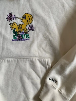 Vit hoodie från Obey  - Vit hoodie från Obey med broderad gul fågel och texten 'LOVE MOVEMENT' på bröstet. Har magficka, huva och OBEY-broderi på ärmen. Mjuk bomullskänsla och avslappnad passform, perfekt för en chill streetwear-look.