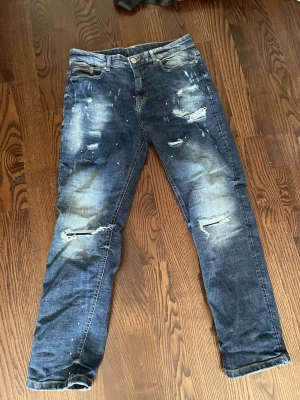 Blå slitna jeans från Supply & Demand - Säljer ett par blå jeans från Supply & Demand med coola slitningar, hål och blekta detaljer. Jeansen har klassisk femficksdesign och knappgylf. Perfekta för dig som gillar en streetig och avslappnad stil.
