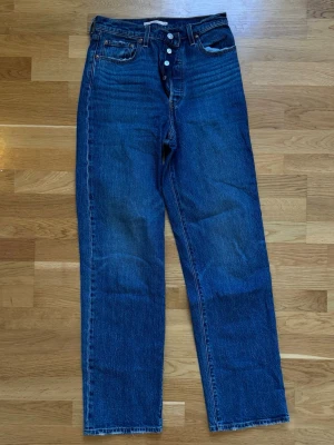 Levi's Ribcage Straight blå jeans - Säljer ett par Levi's Ribcage Straight jeans i klassisk blå denim. Jeansen har hög midja, raka ben och knäppning med knappar framtill. Ikonisk femficksmodell i 100% bomull som ger en tidlös och cool look.